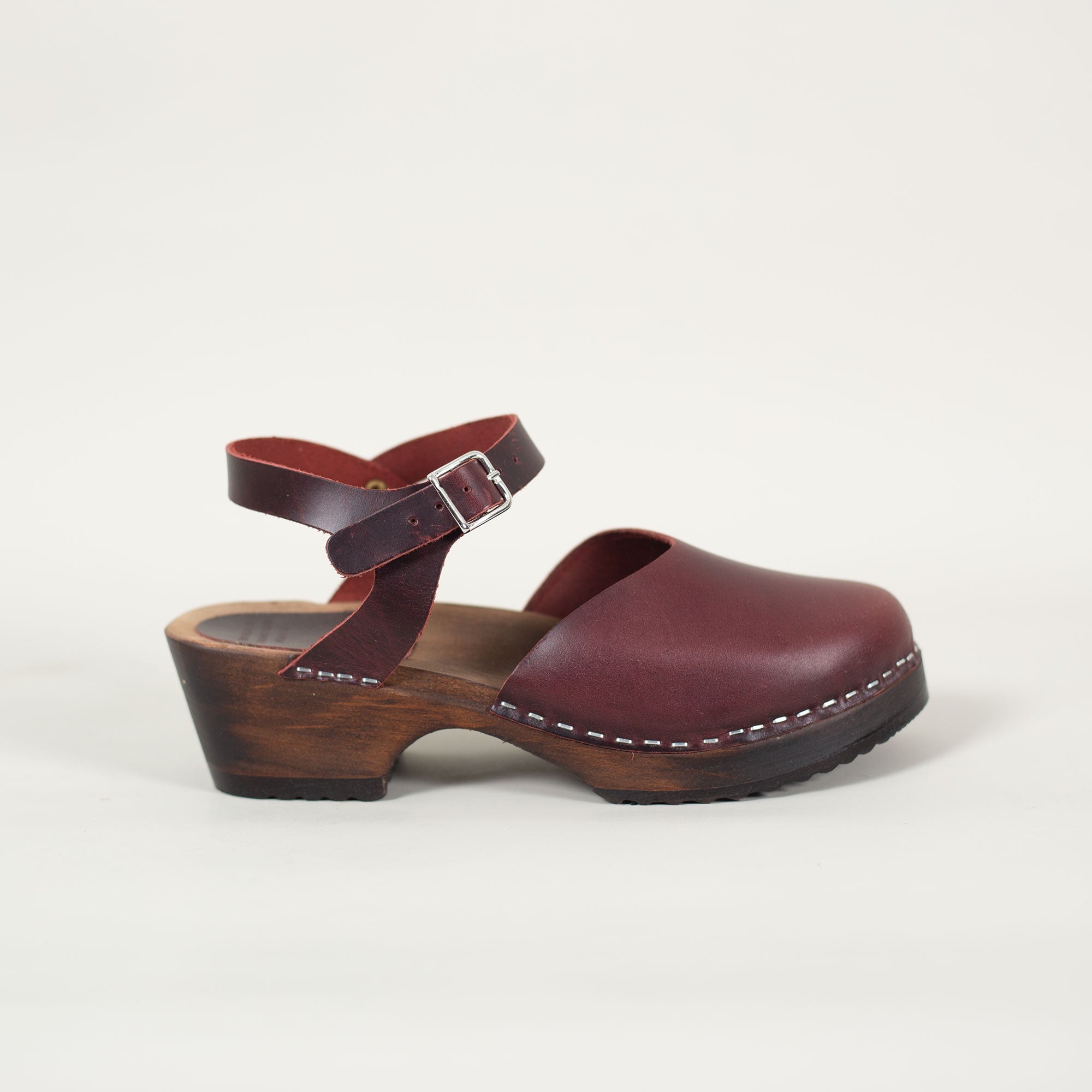 A bordeaux closed-toe clog sandal with an adjustable ankle strap and a low heel, made with a bordeaux pull up leather upper and brown stained & varnished alderwood.